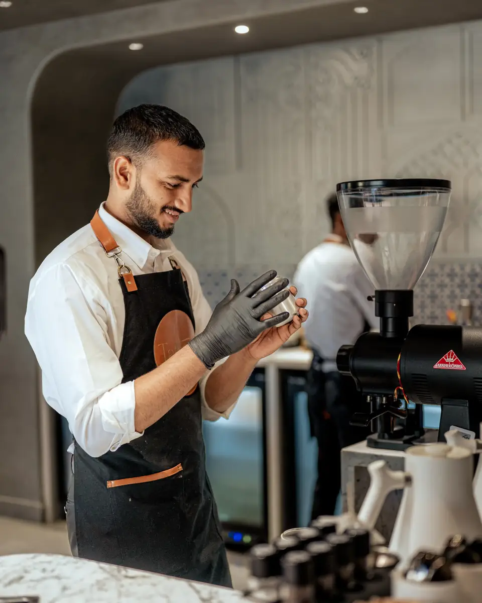 A barista adjusting black gloves beside the grinder, under an arched niche