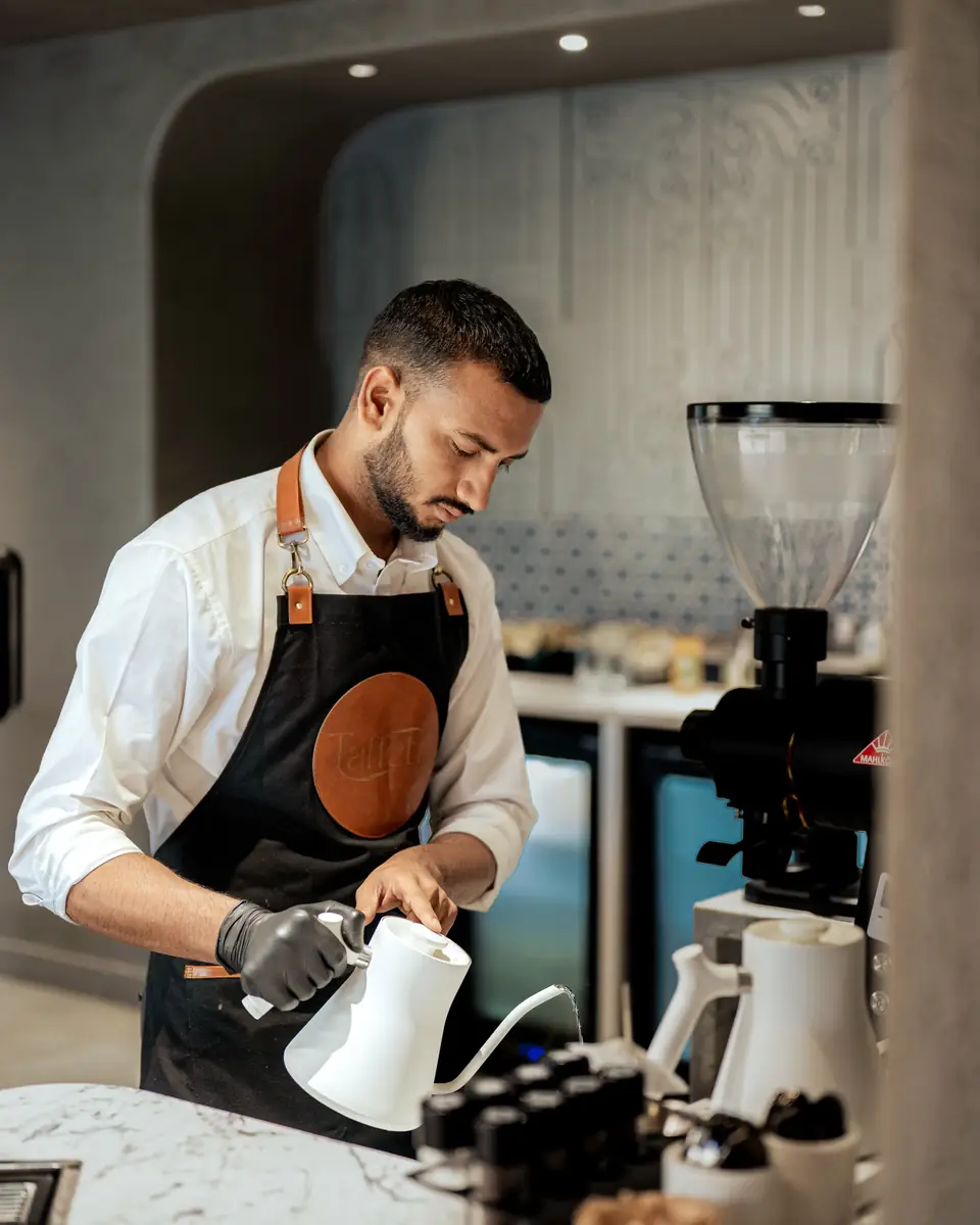 A barista pouring from a gooseneck kettle beside the grinder