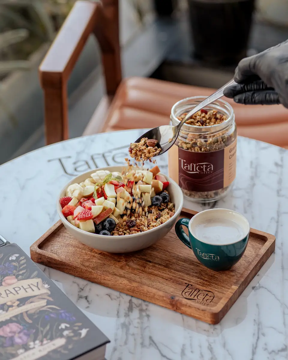 Granola being spooned over a fruit and yoghurt bowl next to a cup of milk