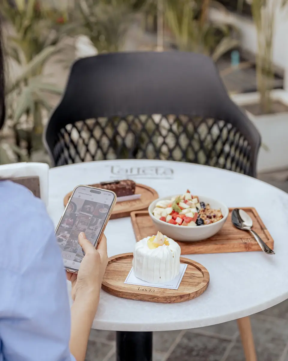 A guest on a phone at a marble terrace table with a fruit bowl and a cake