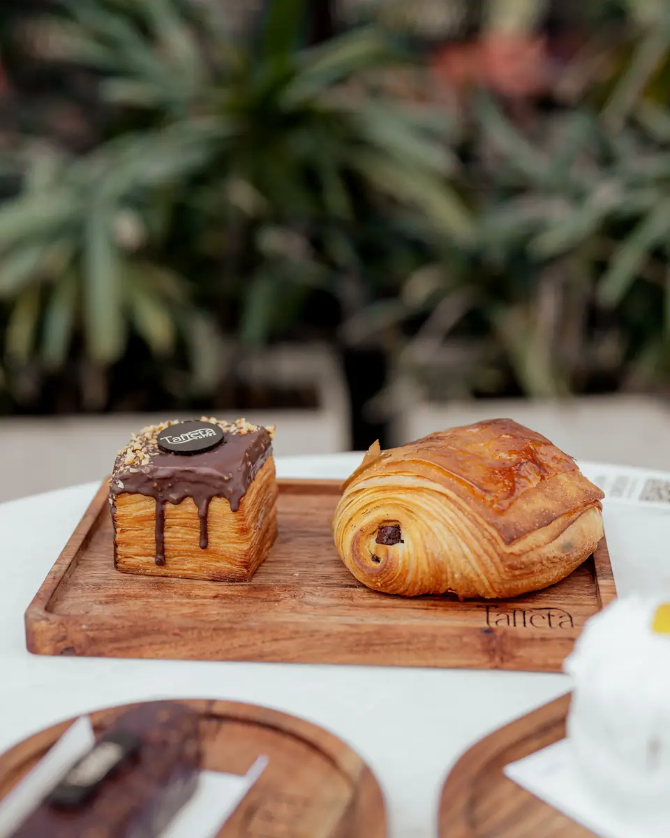 A drip-glazed chocolate pastry beside a pain au chocolat on a wooden board