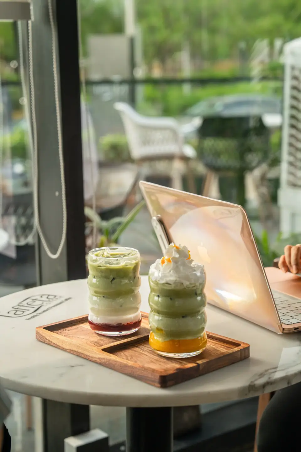 Two layered drinks on a tray at a window table, next to a laptop