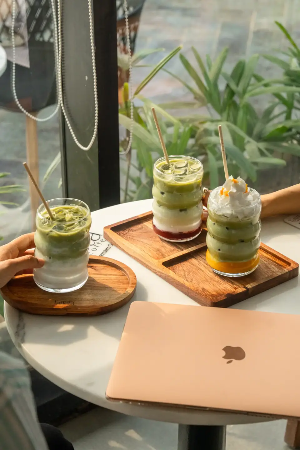 Three layered iced drinks on a wooden tray beside a laptop at a window table