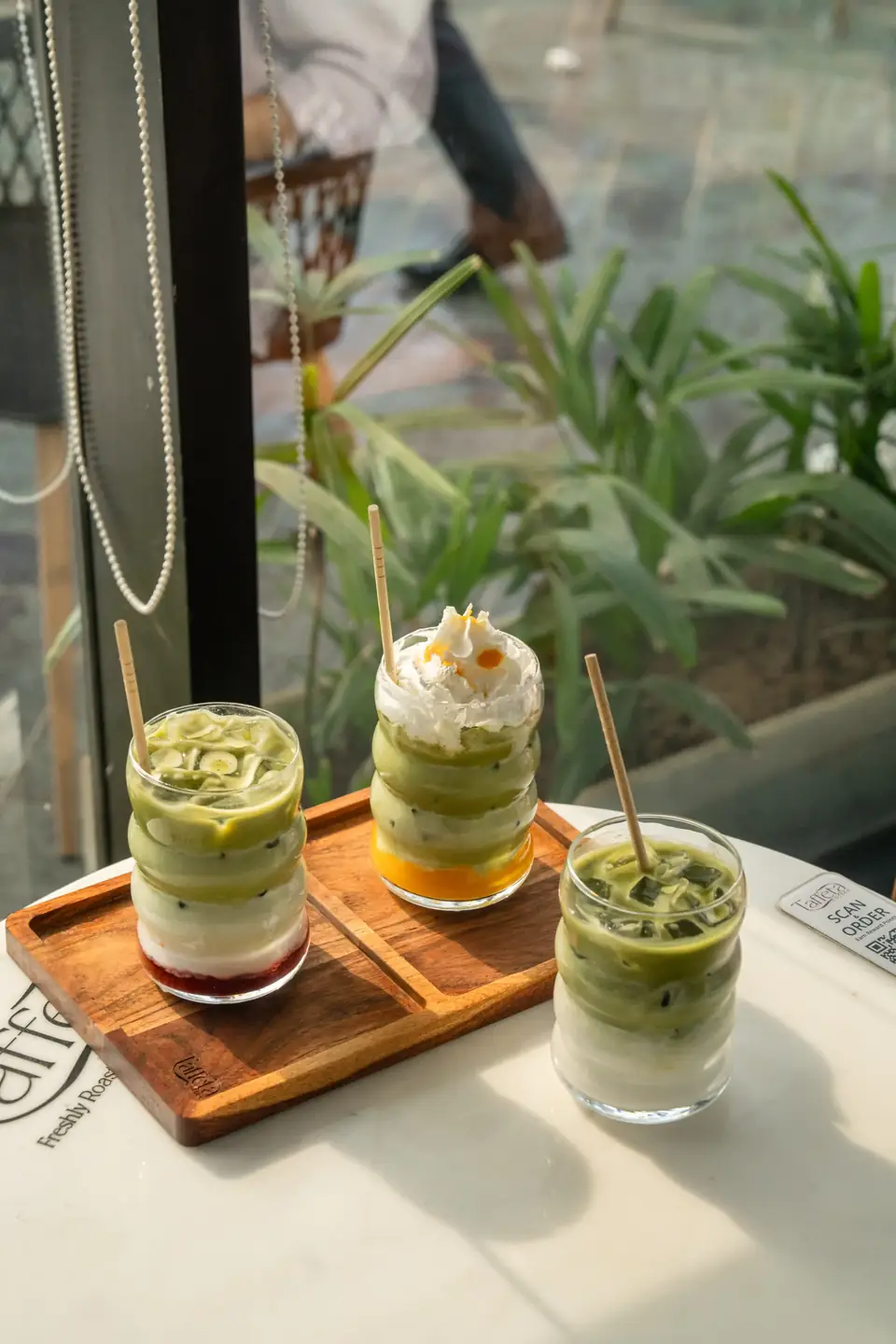 Three layered iced drinks on a wooden tray by the window, plants outside