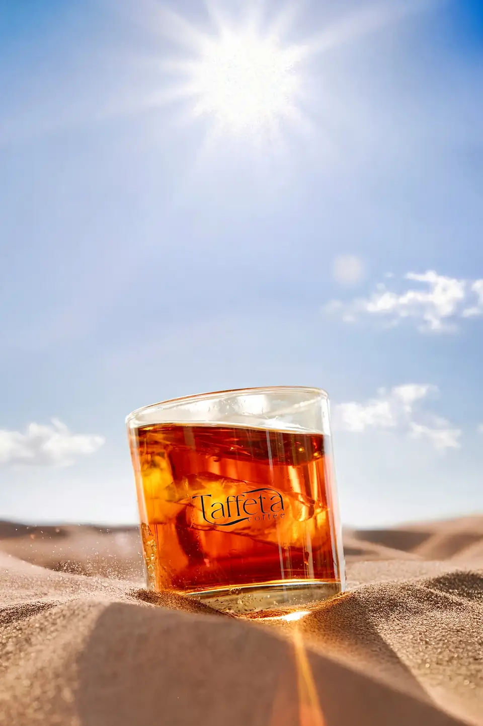 A glass of iced coffee with the Taffeta logo standing on sand under a bright sun