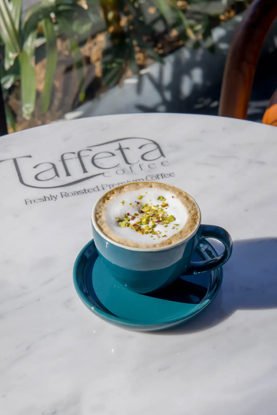 A pistachio-topped cappuccino in a teal cup on a Taffeta marble table